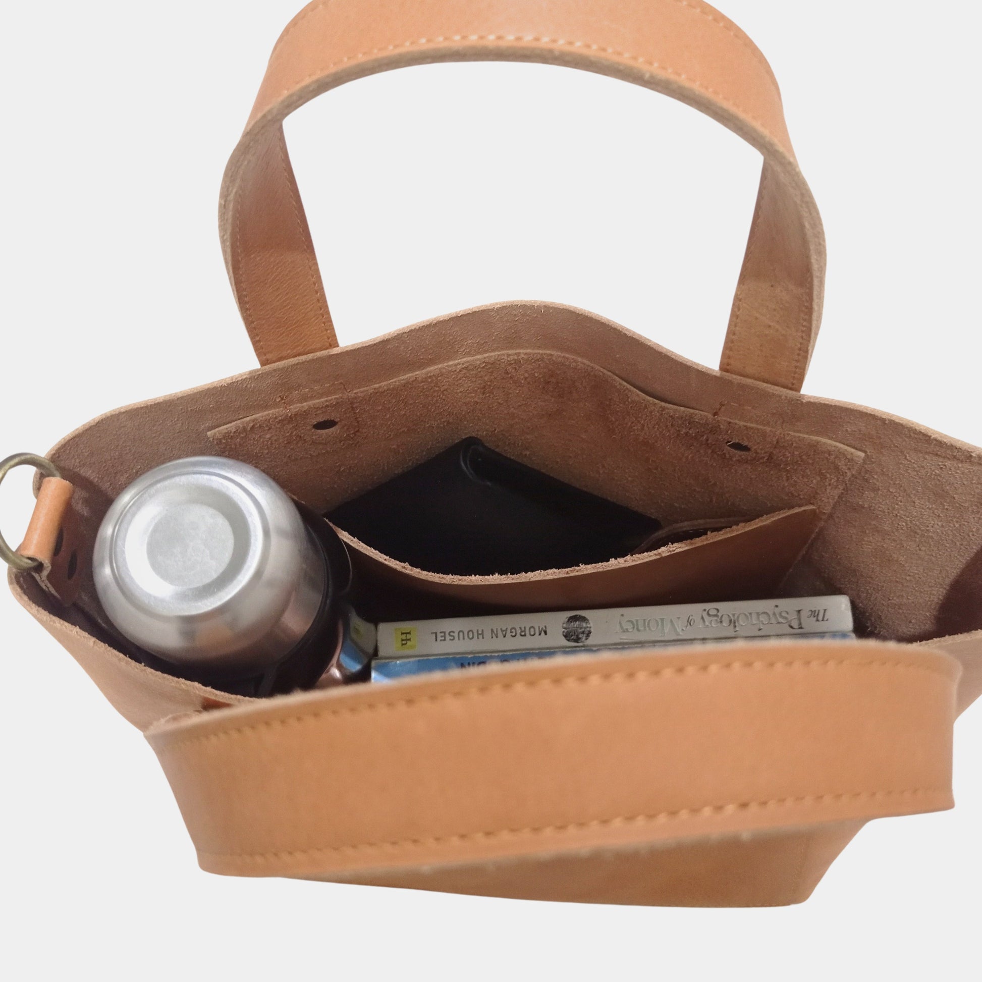 Inside of unlined brown leather tote showcasing hanging pocket with black wallet, books and water bottle 